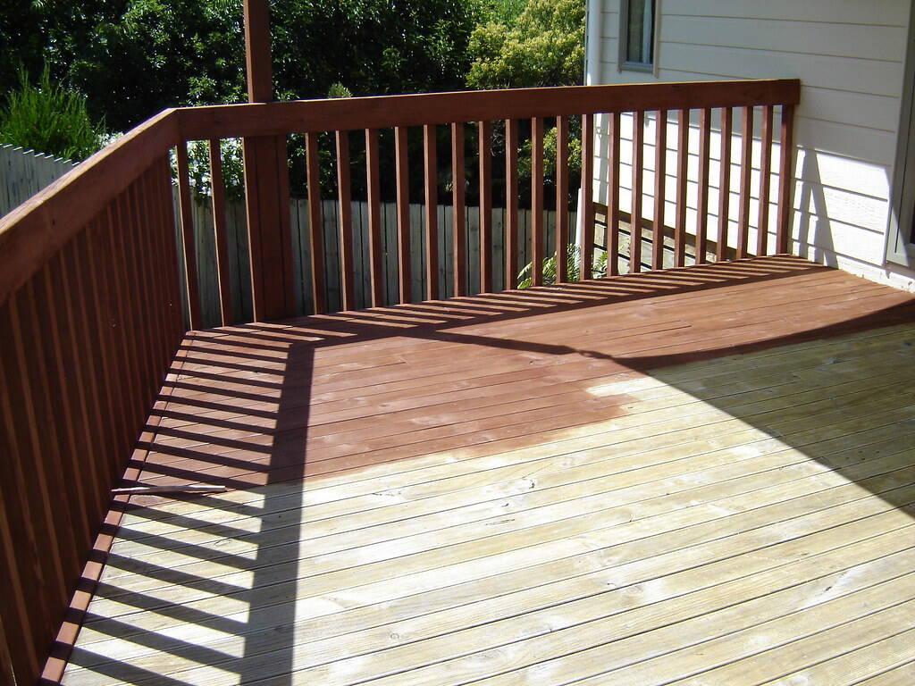 A backyard wood deck mid-project, half the boards freshly stained and half still bare.