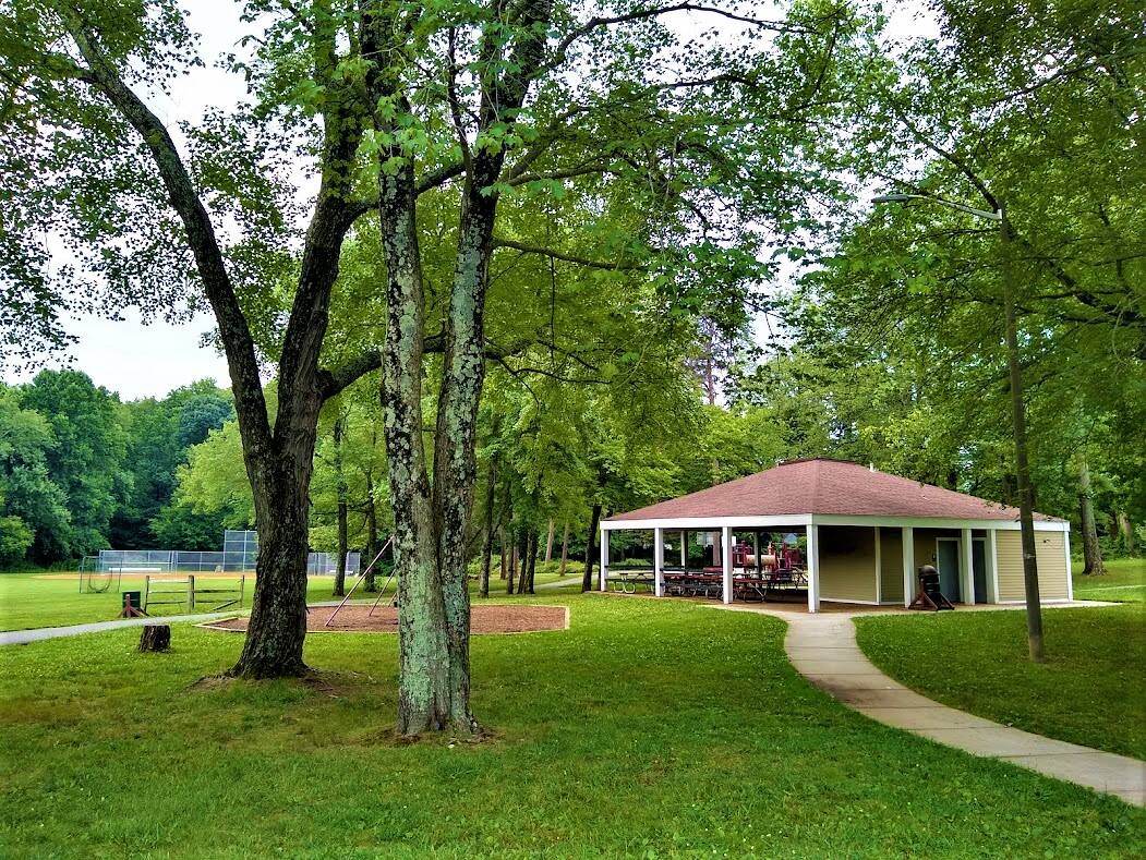 A village park in full summer green: an open-sided pavilion with picnic tables, a winding path, and a ball field beyond.