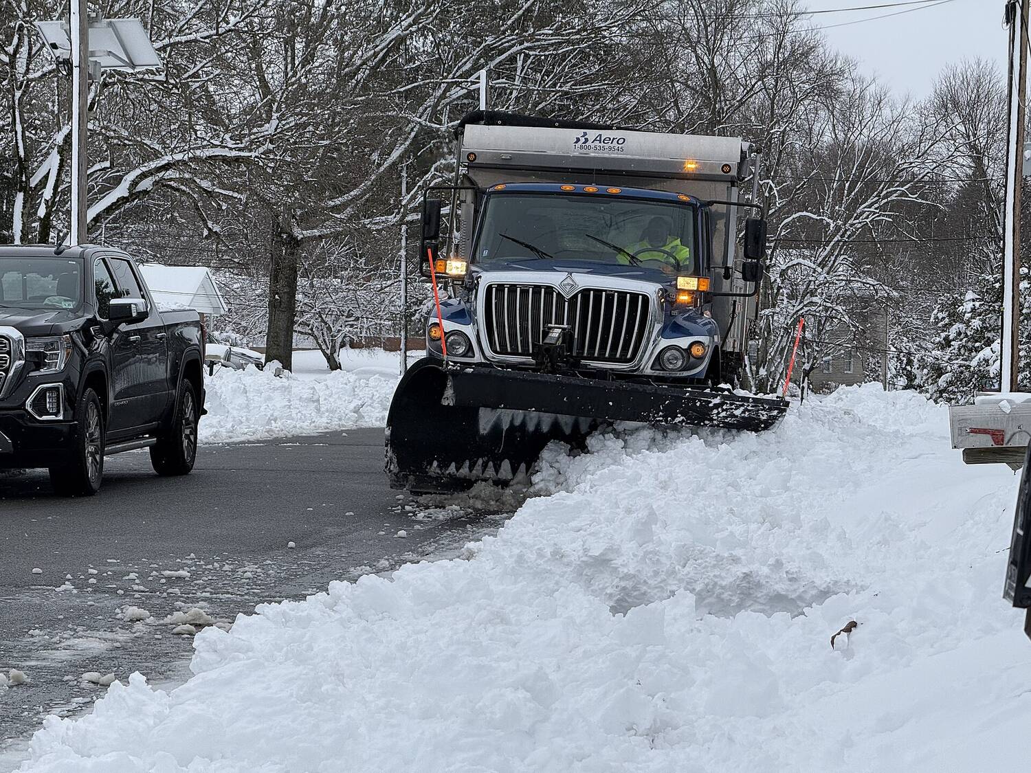 A municipal snow plow truck clearing a residential street after a winter storm, snow banked on both sides.