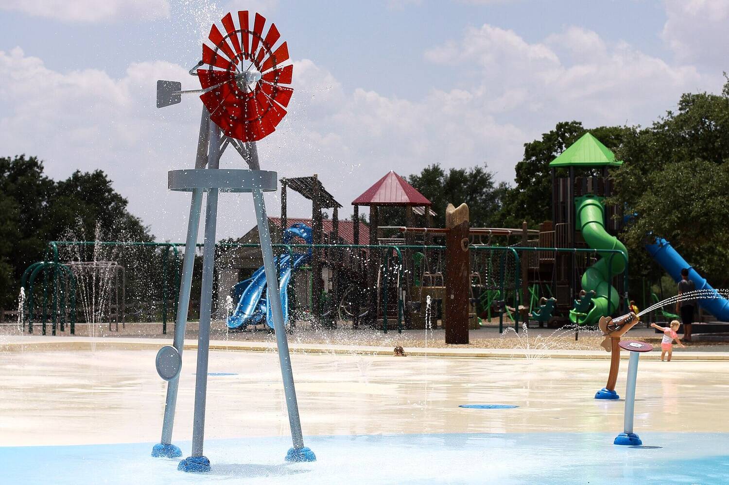 A splash pad with a red windmill-shaped water feature spraying water, playground in the background.