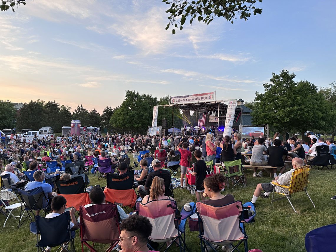 A large outdoor community event in Gilberts, with families seated in lawn chairs facing a stage at dusk.