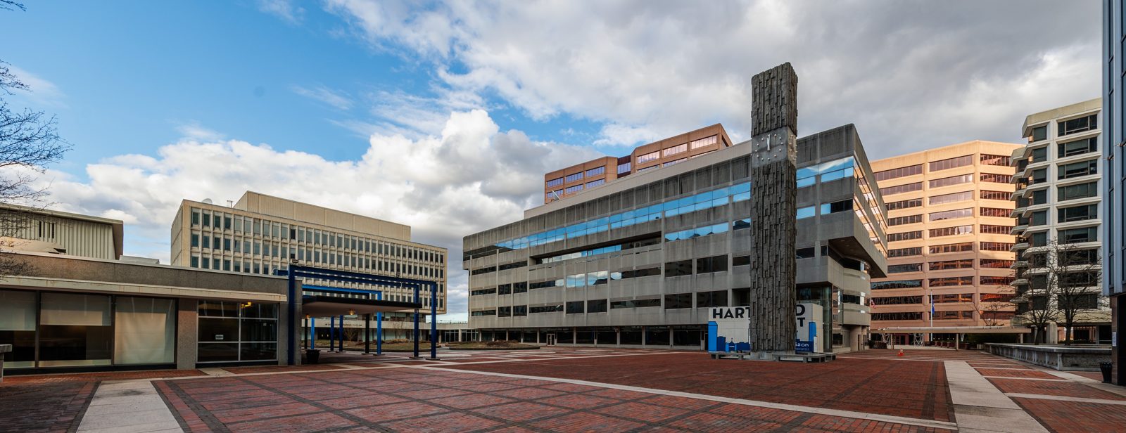 A wide view across Constitution Plaza in Hartford, its brick-paved deck running between mid-century office towers, with Masao Kinoshita's stone clock tower standing at the centre.