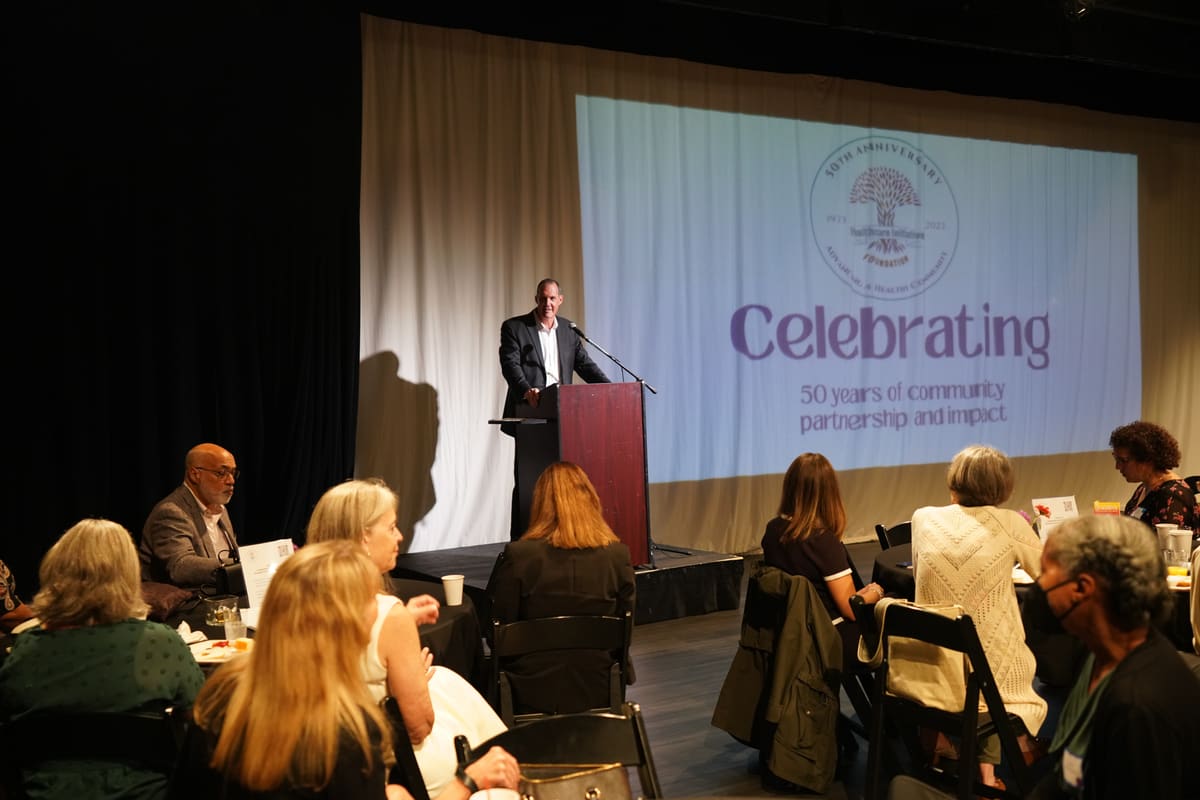 Opening remarks at the podium under the "Celebrating 50 years of community partnership and impact" screen at HIF's 50th Anniversary breakfast