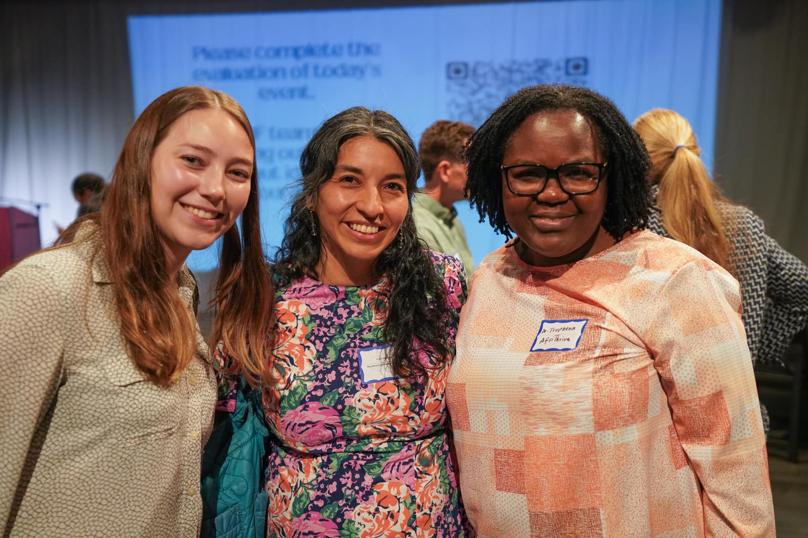 Three women from the American Heart Association and AfriThrive smiling together in front of the event screen at HIF's 50th Anniversary breakfast
