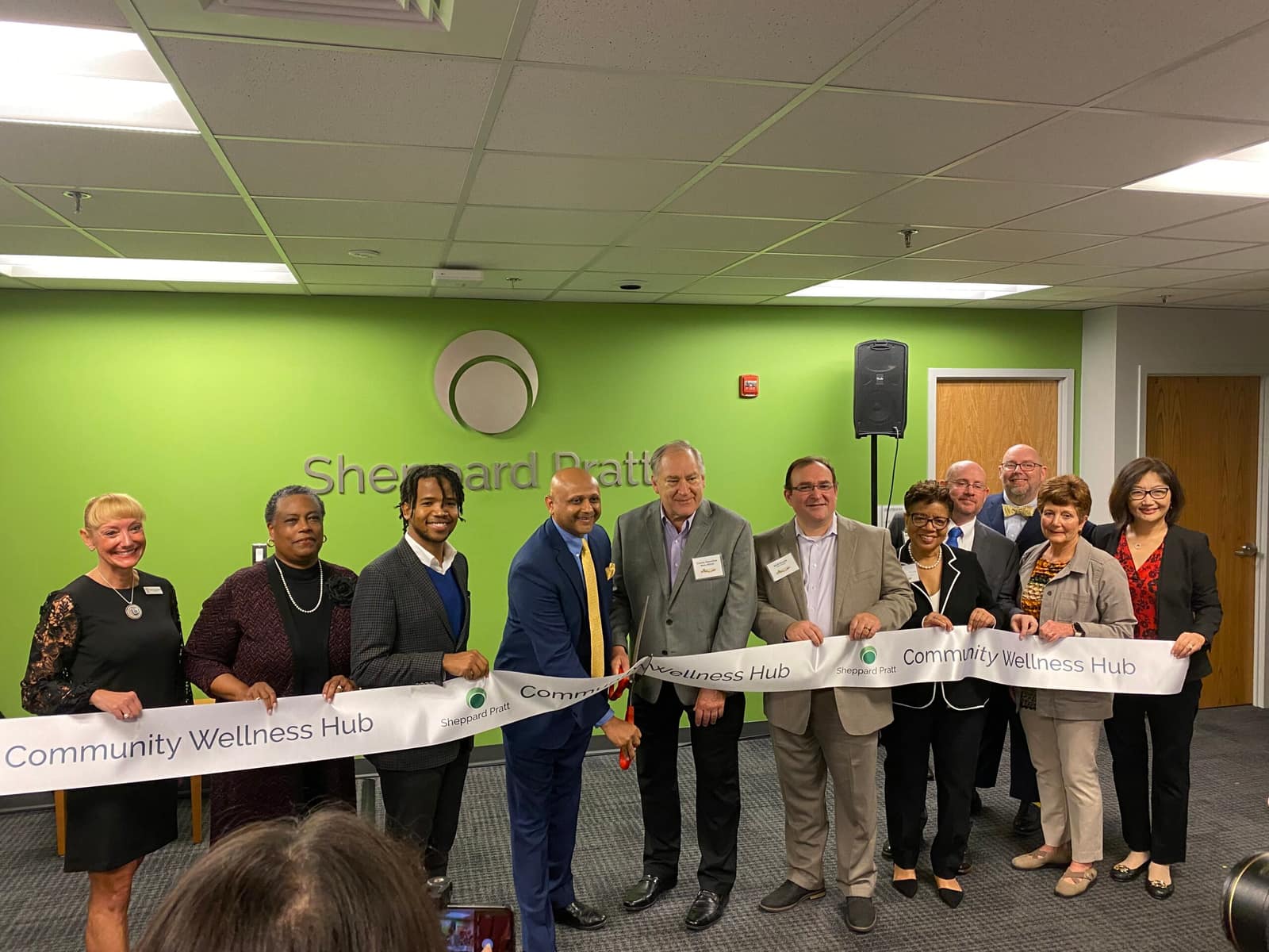 The ribbon cutting at Sheppard Pratt's Community Wellness Hub, eleven people holding the ribbon in front of the green Hub wall