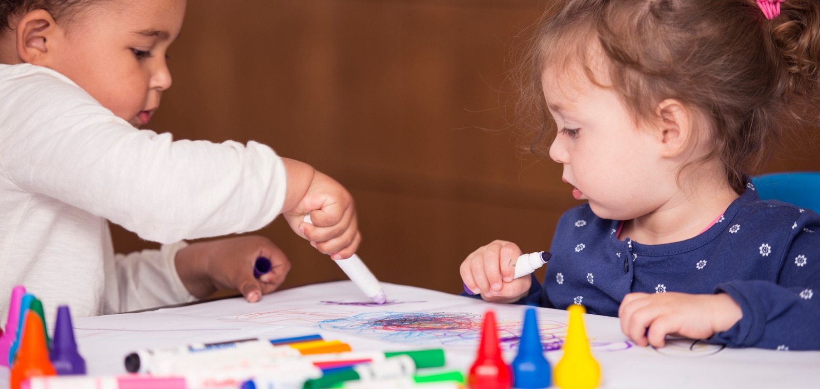 Two toddlers coloring at a table with markers in a child care room