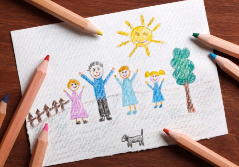 A child's crayon drawing of a family and a house, photographed with colored pencils beside it