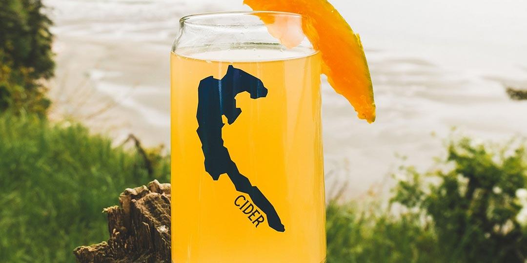 A glass of Camano Ciders' cider with the island silhouette on it, poured against the beach grass