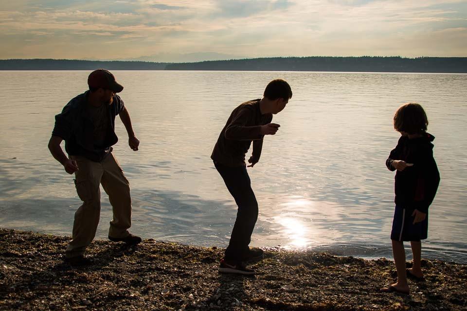 Three silhouetted figures skipping stones off the Camano shore in evening light