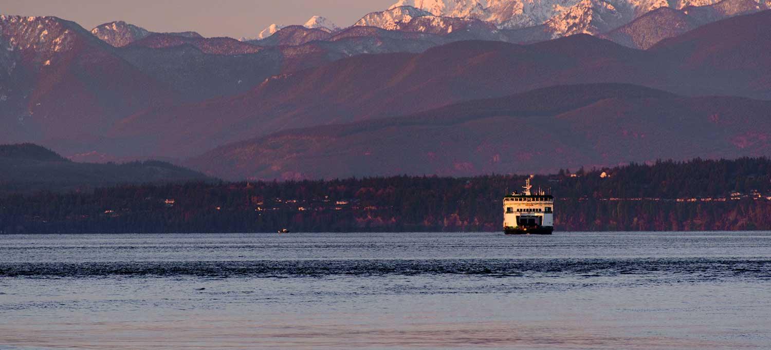 A Washington State ferry crossing at dusk, the Olympic Mountains layered pink behind it