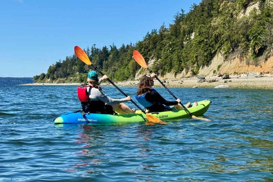 Two paddlers in a green tandem kayak under the wooded bluffs of Camano Island
