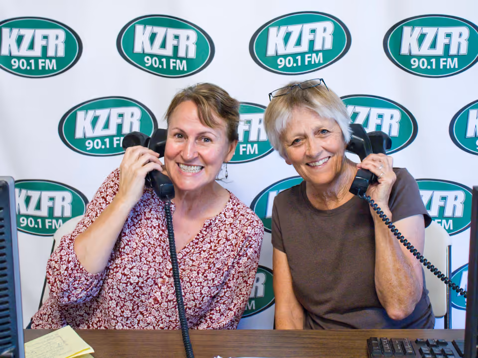 Two smiling volunteers hold telephone handsets to their ears at a desk, ready to take pledges, in front of a KZFR 90.1 FM banner