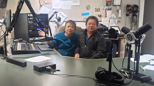 Two Tshua Hmoob hosts sit side by side at the desk in the KZFR studio, surrounded by microphones and mixing equipment