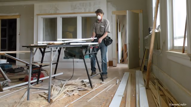 Man in a dust mask and tool belt cutting trim at a table saw in a room under renovation, wood scraps on the floor