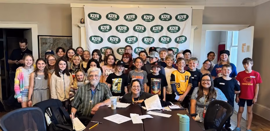 Group of about thirty Chico Country Day School 5th graders and adults posing in front of a KZFR 90.1 FM banner, with two people seated at a table of papers