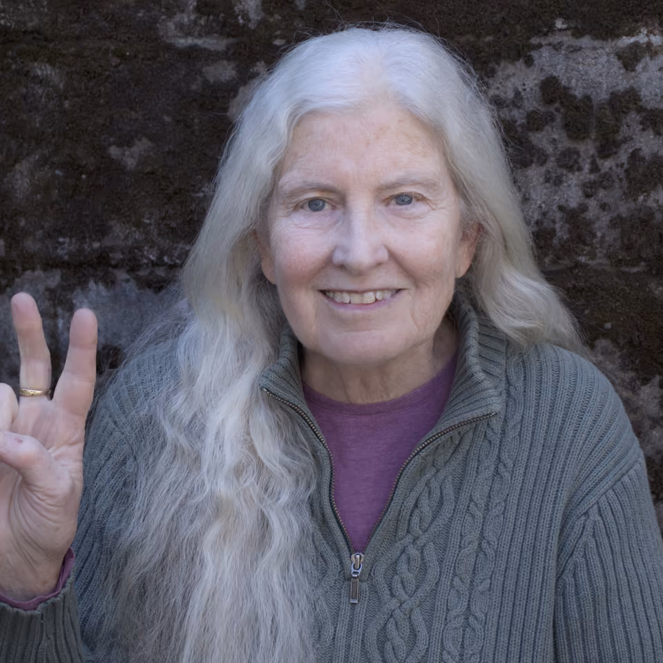 Smiling woman with long gray hair and a green zip cardigan holds up a peace sign against a mossy stone wall