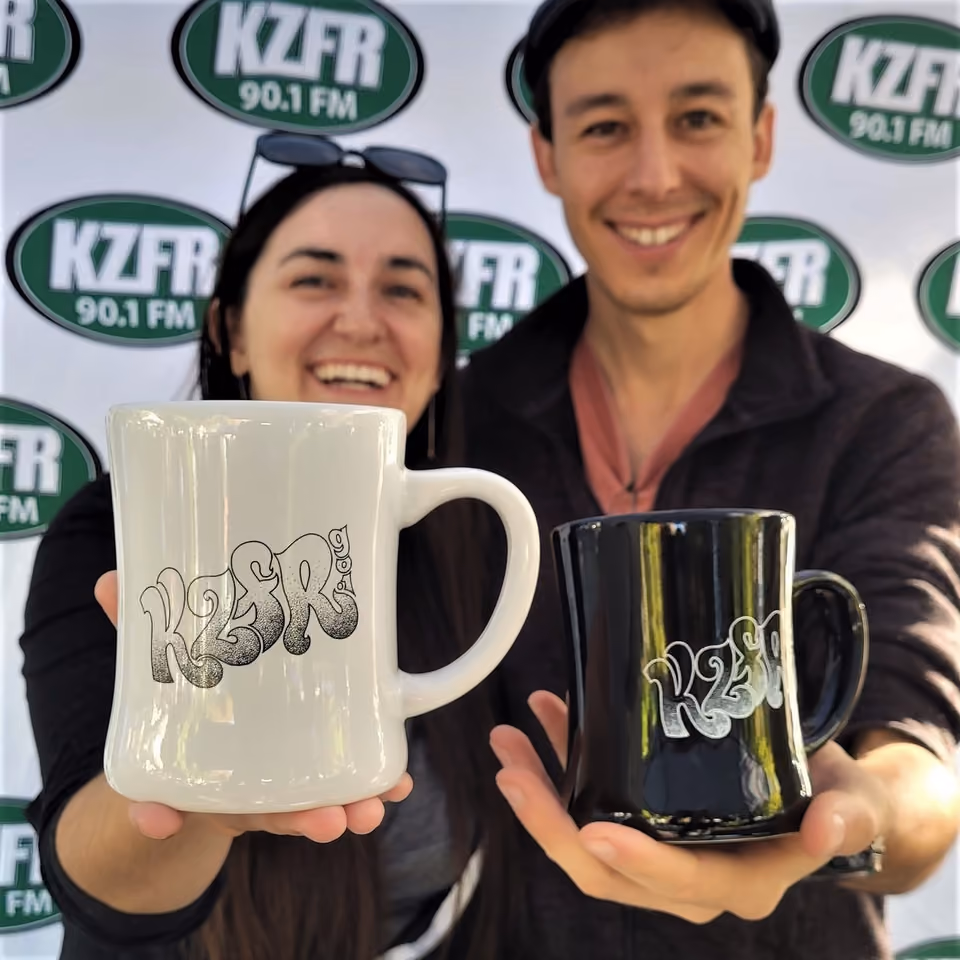 Two smiling volunteers in front of a KZFR 90.1 FM banner hold up white and black KZFR logo coffee mugs, thank-you gifts</br>

Correction: reply with the alt text only.

Two smiling people in front of a KZFR 90.1 FM banner hold up white and black KZFR logo coffee mugs