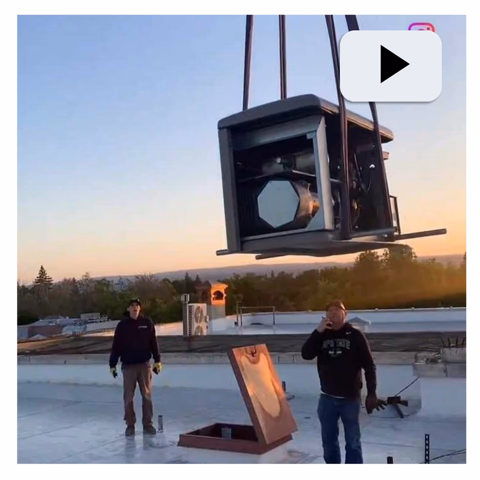 Video still of a generator hoisted by crane above a rooftop at sunset, with two workers watching from below