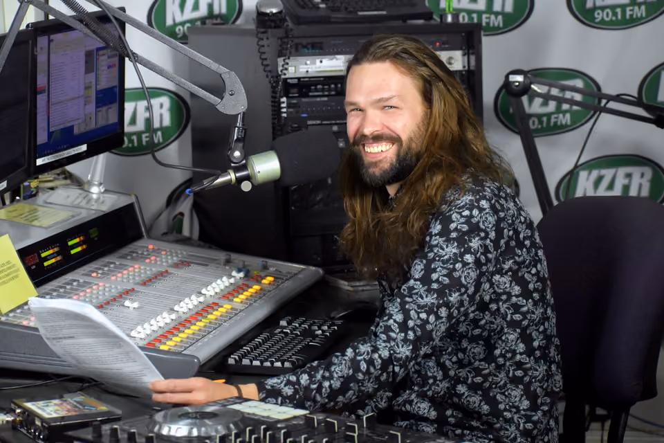 Smiling long-haired, bearded host seated at the mixing board in the KZFR studio, holding a paper script
