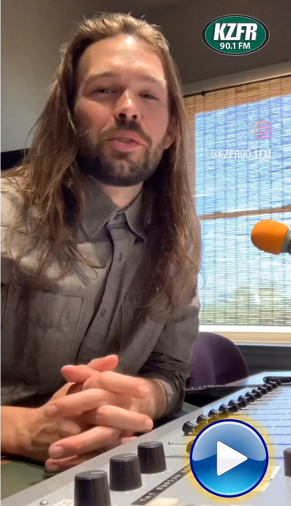 Video still of a long-haired, bearded man in a gray shirt speaking at the KZFR studio mixing board, with a play button overlay