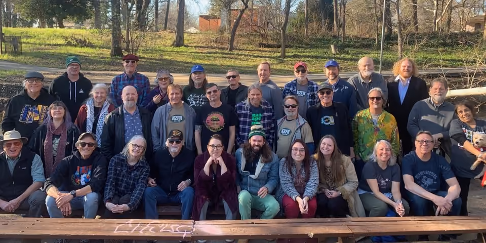 Group photo of about two dozen smiling KZFR volunteers posed outdoors in rows on and behind a wooden bench, many in station T-shirts