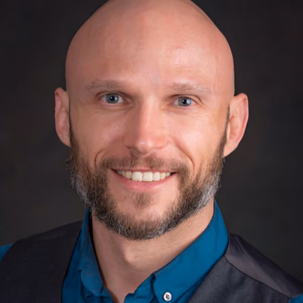 Headshot of a smiling bald man with a graying beard, wearing a teal collared shirt and dark vest, against a dark background