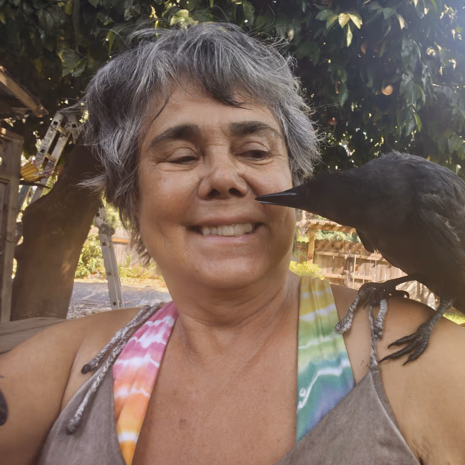 Smiling woman with short gray hair outdoors under a tree, a black crow perched on her shoulder with its beak near her cheek