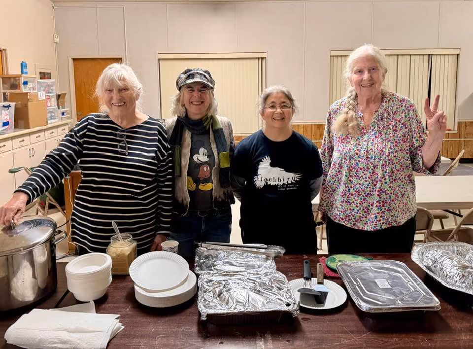 Four smiling KZFR programmers stand behind a table of paper plates, serving pots and foil trays after serving a meal at Safe Space