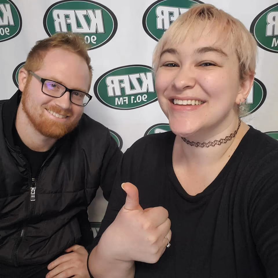 Two smiling people pose in front of a KZFR 90.1 FM banner, one giving a thumbs up