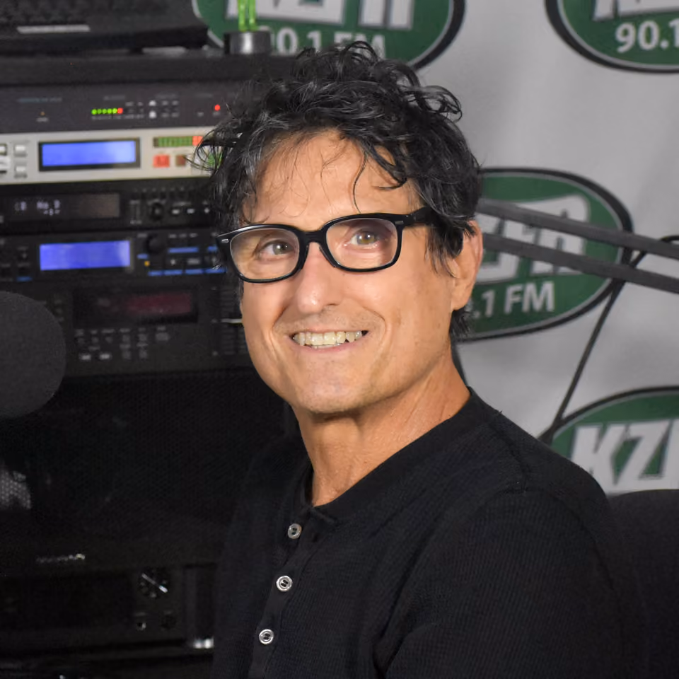 Smiling volunteer with dark curly hair and black glasses sits in the KZFR studio before audio equipment and a station logo backdrop