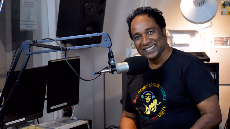 Smiling Sanjay Dev, host of Devastation Sounds, sits at the studio microphone in a black reggae lion T-shirt