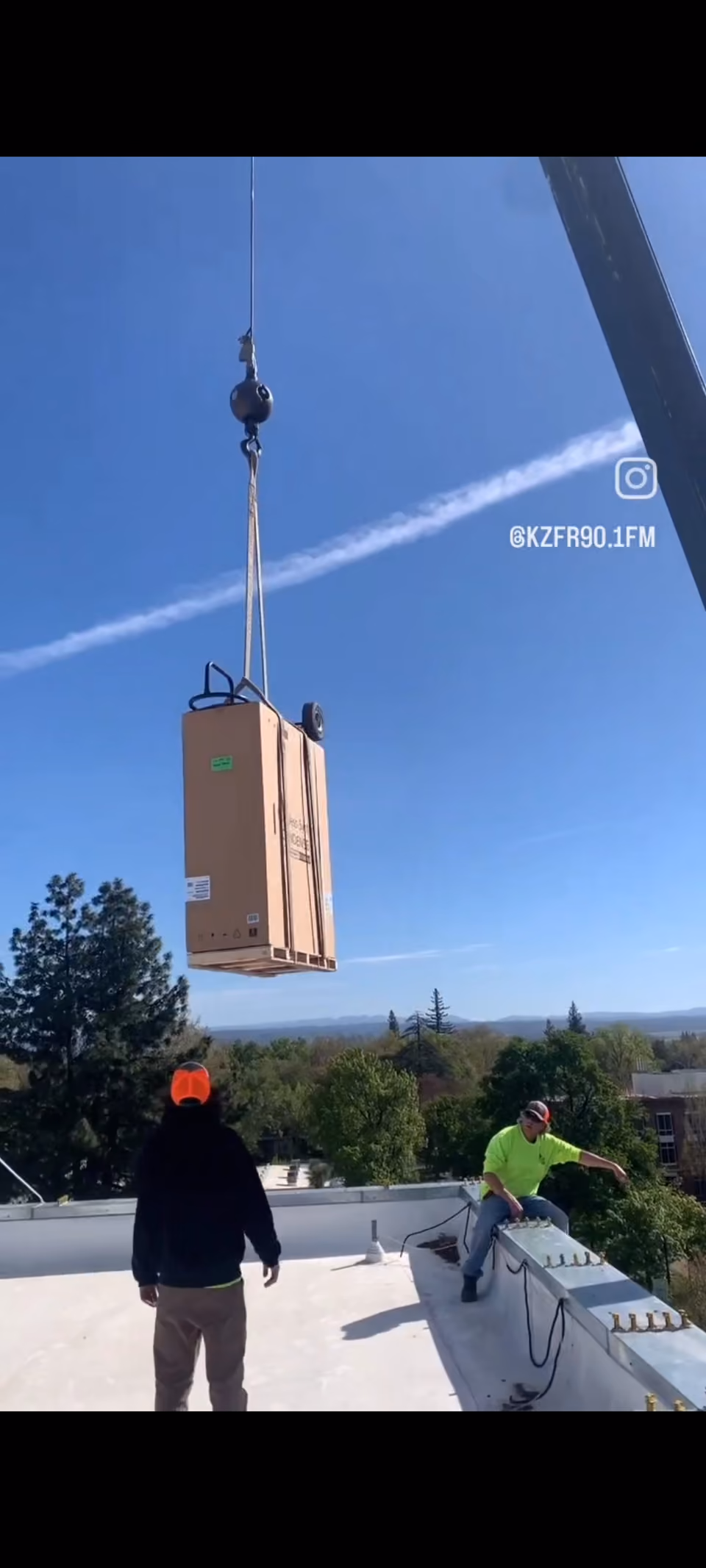 Crane lifting a boxed HVAC unit above a rooftop while two workers guide it, under a clear blue sky