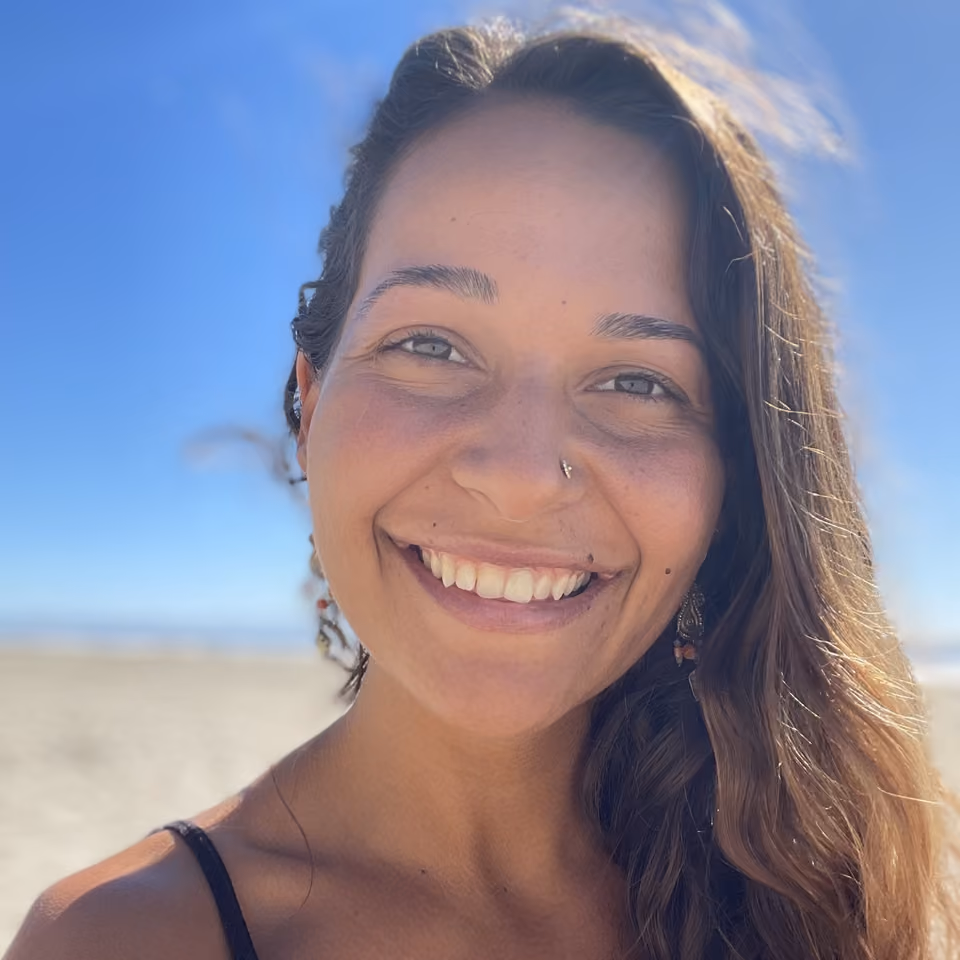 Smiling woman with long brown hair and a nose stud takes a selfie on a sunny beach under a blue sky