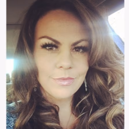 Close-up selfie of a woman with long wavy brown hair, dramatic lashes and drop earrings, sunlight streaming in from a car window