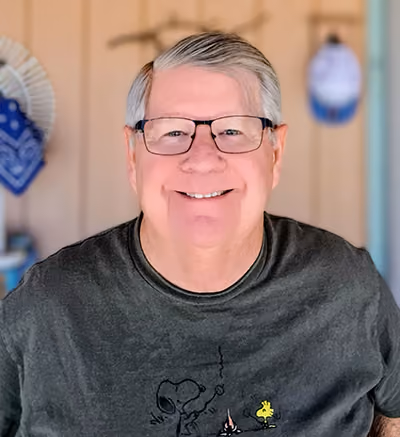 Smiling older man with gray hair and glasses, wearing a dark gray Snoopy and Woodstock t-shirt, a board member portrait