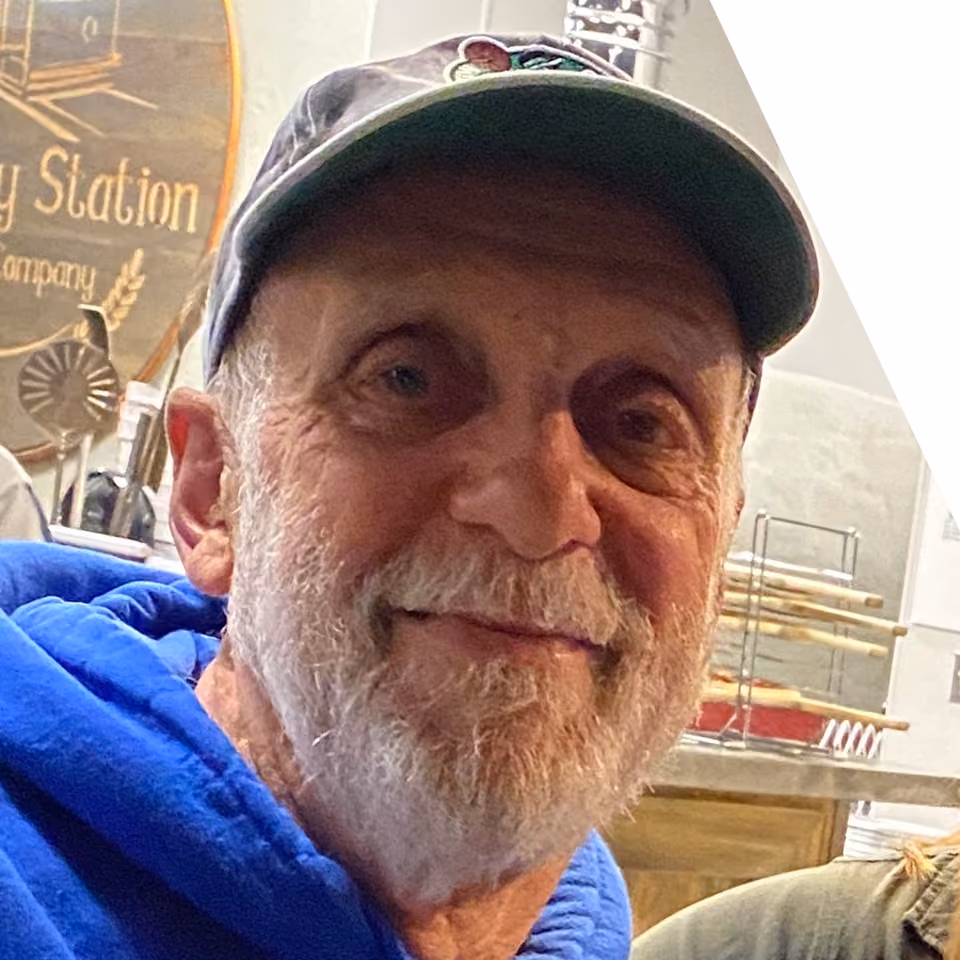 Smiling older man with a white beard, wearing a gray cap and blue hoodie, standing in a restaurant or bakery