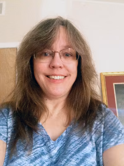 Smiling woman with long brown hair, bangs and wire-rimmed glasses, wearing a blue patterned top, in a home interior