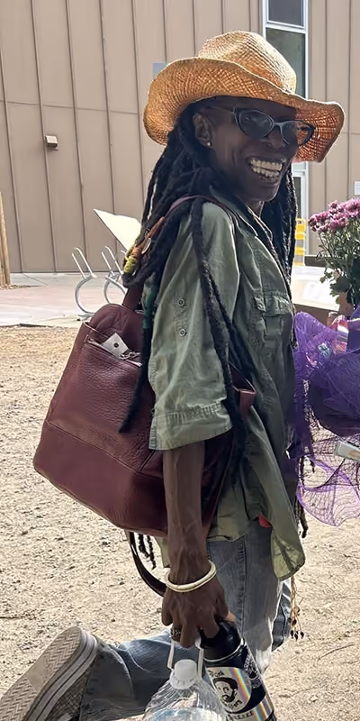 Smiling woman in a straw hat and glasses, with long locs, a green shirt and a maroon shoulder bag, standing outdoors near purple flowers