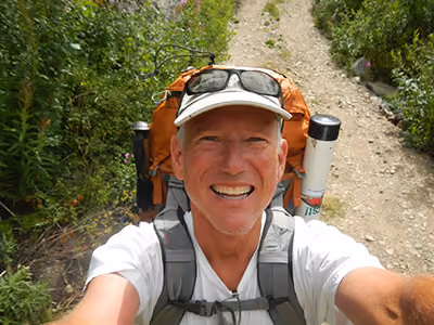 Smiling man in a white cap and sunglasses takes a selfie on a gravel trail, wearing an orange hiking backpack