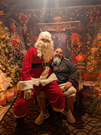 Person in a Santa Claus costume sits beside a smiling bearded man in a festive holiday setting with decorated trees and gifts