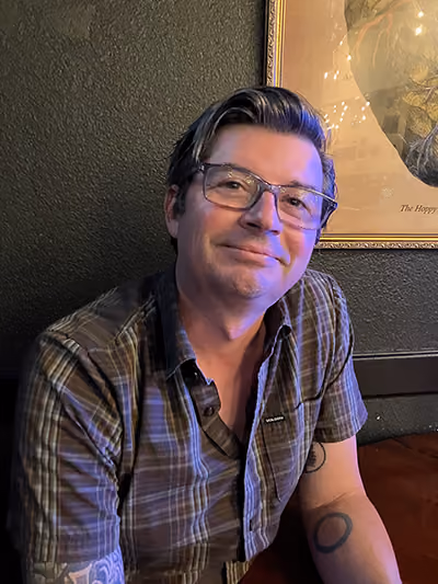 Smiling man with glasses and swept-back graying hair, wearing a plaid short-sleeve shirt, seated in front of a framed picture, with tattoos on his arms