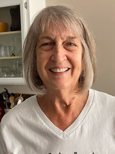 Smiling older woman with shoulder-length gray hair and a white V-neck shirt, standing in a kitchen with a glass-front cabinet behind her