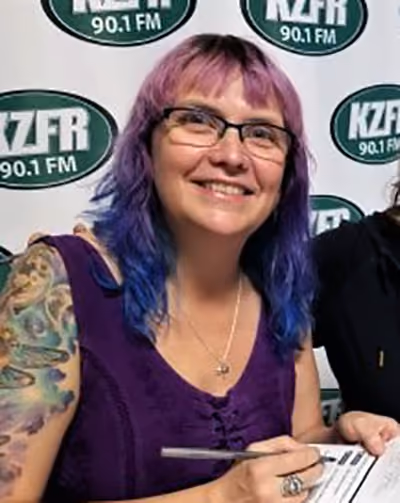 Smiling person with purple and blue hair and glasses, holding a pen in front of a KZFR 90.1 FM back