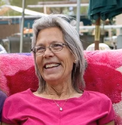 Smiling woman with gray shoulder-length hair and glasses, wearing a pink top and pendant necklace, seated outdoors against a pink fuzzy chair