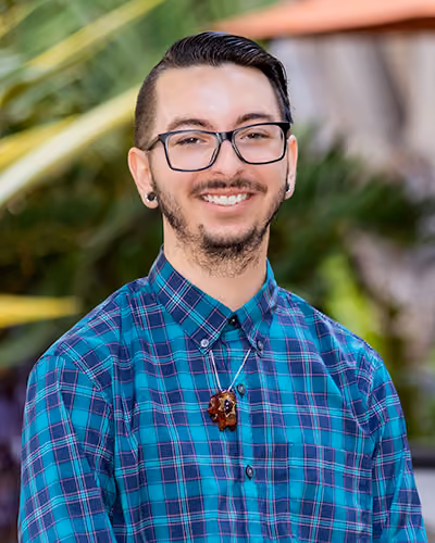 Headshot of a smiling board member with glasses, a goatee and slicked-back dark hair, wearing a teal plaid shirt and a wooden pendant necklace, outdoors in front of green foliage