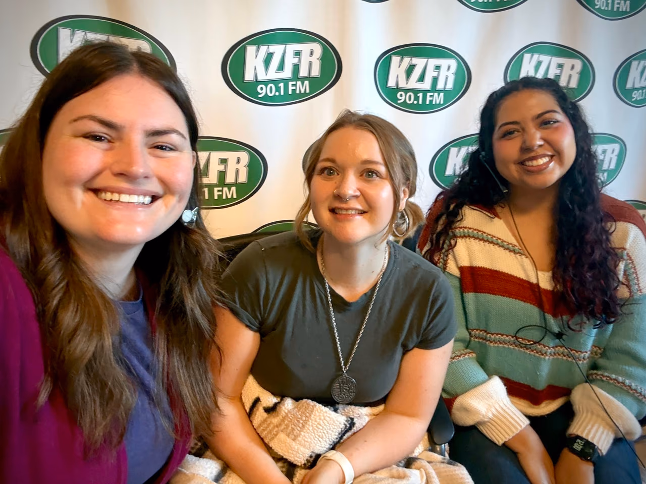 Three volunteers from Catalyst Domestic Violence Services smiling in front of the KZFR backdrop during the pledge drive.