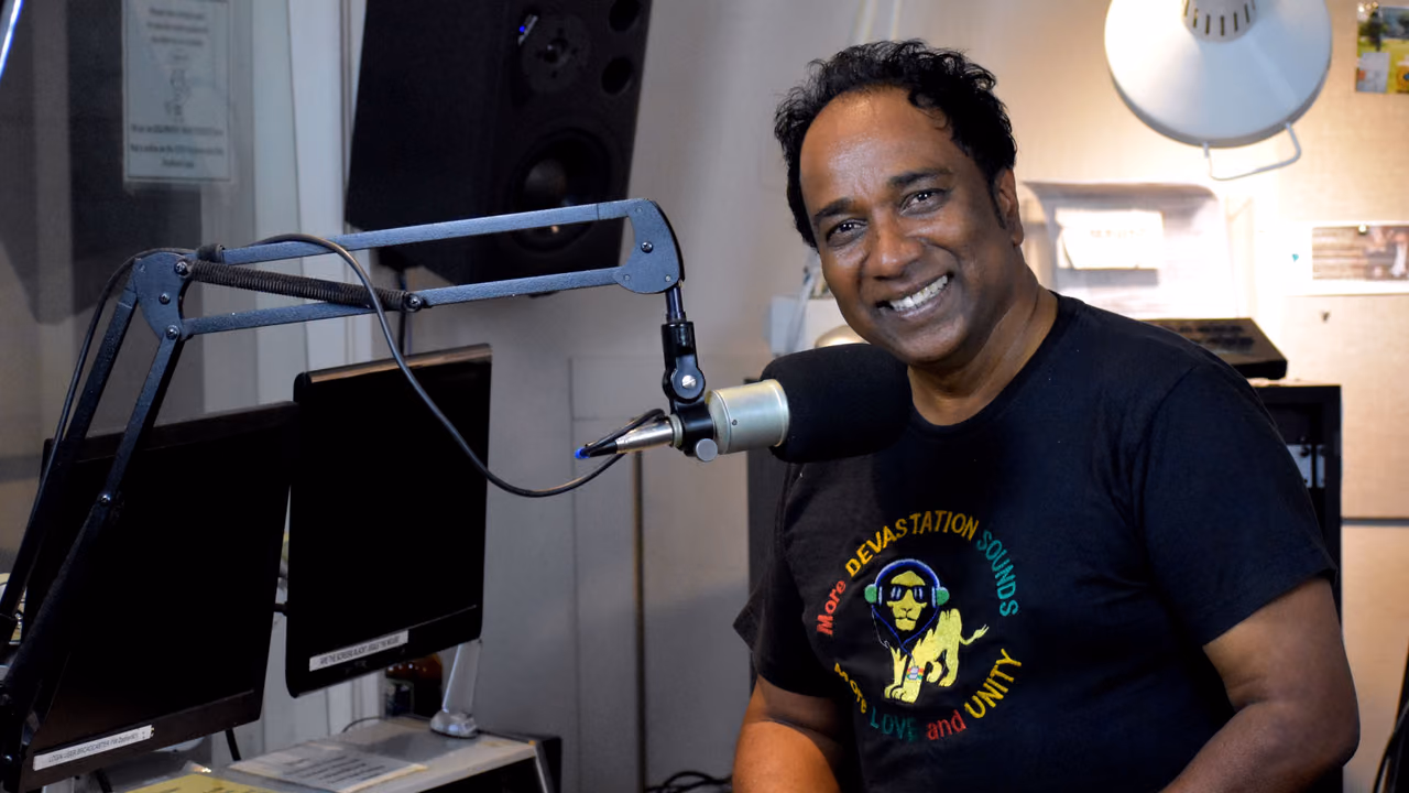Sanjay Dev smiling at the KZFR studio board in a KZFR T-shirt, a monitor and the mixing board beside him.