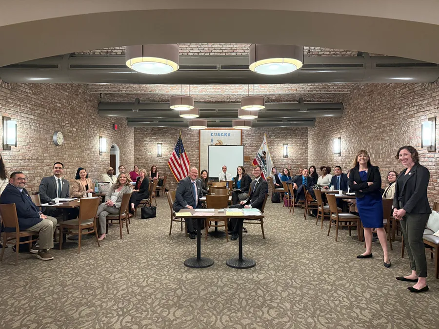 About thirty people in a brick-walled meeting room turn toward the camera from tables and chairs, with a United States flag and a California state flag standing behind them.
