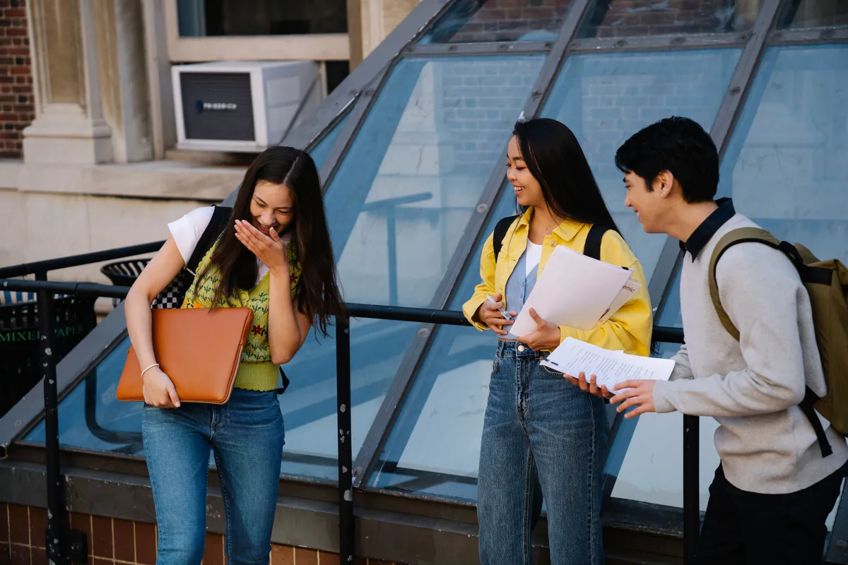 Three students sharing a laugh outside a campus building, holding notebooks and a laptop.