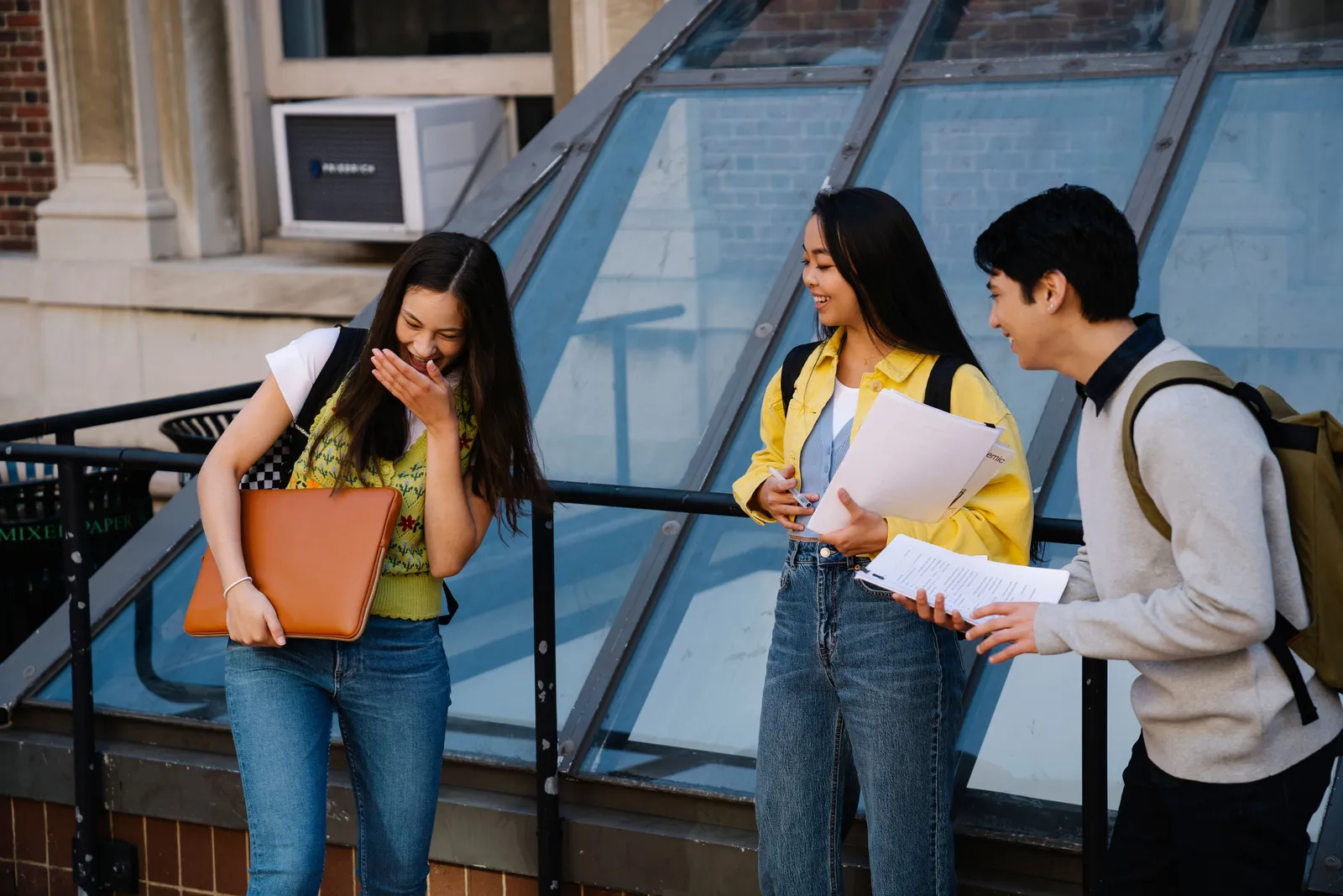 Three students sharing a laugh outside a campus building, holding notebooks and a laptop.